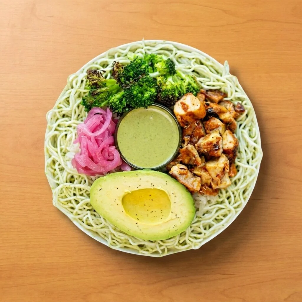 Avocado Citrus Bol: Jasmine rice and pesto noodles paired with garlic broccoli, lemon chicken, pickled red onions, and a fresh avocado half, drizzled with herb pesto.
